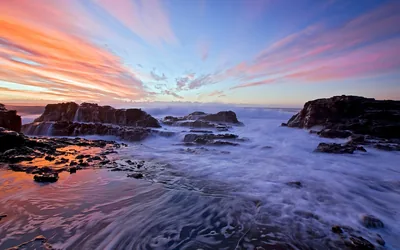 Pink cloud streaks fan across a dawn sky while long exposure swirls the receding foam into rings between the black rocks below.