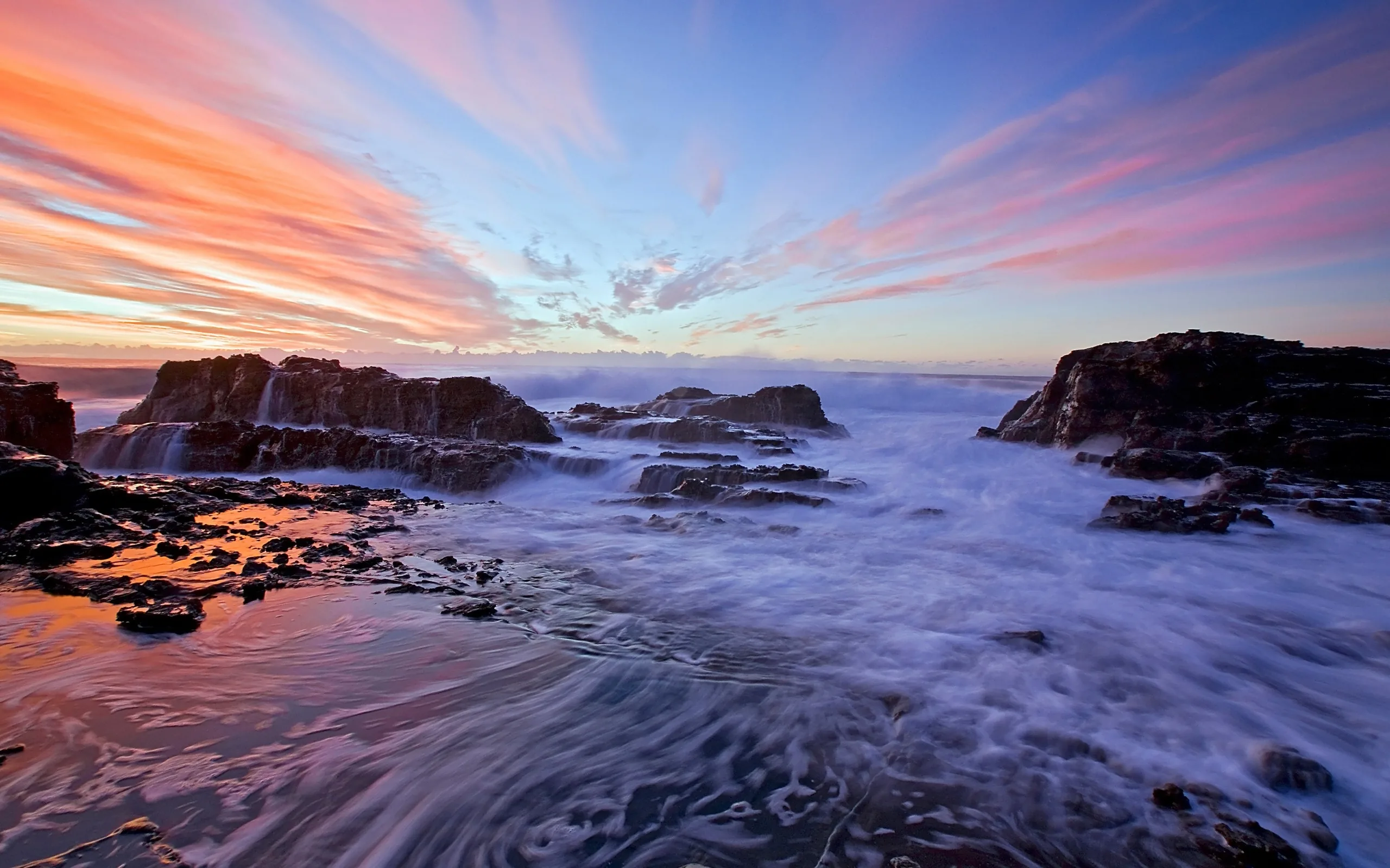 Pink cloud streaks fan across a dawn sky while long exposure swirls the receding foam into rings between the black rocks below.