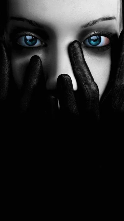 Two black gloved hands cover the lower half of a pale face, leaving only bright blue eyes and dark brows lit against the shadow.