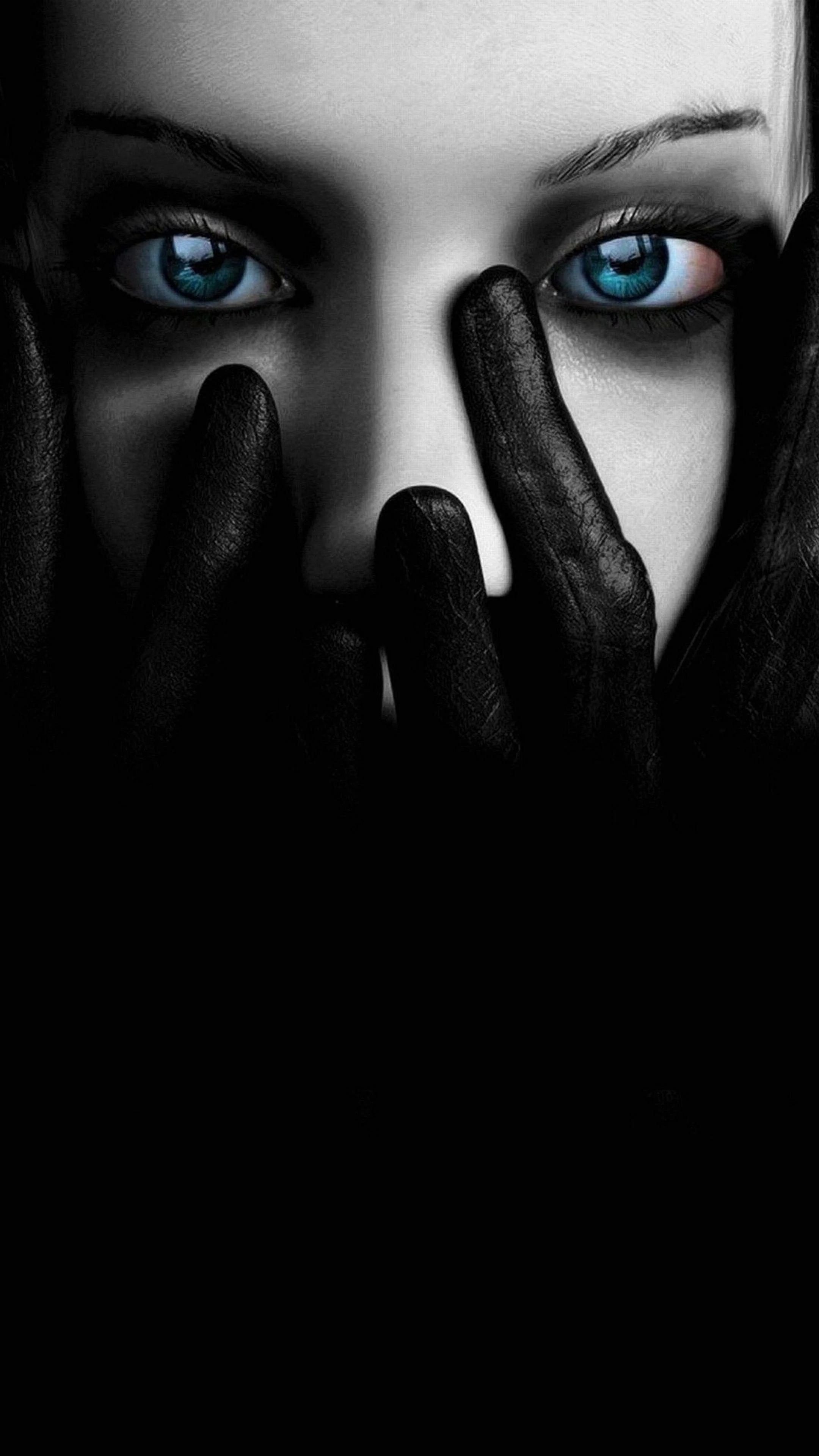 Two black gloved hands cover the lower half of a pale face, leaving only bright blue eyes and dark brows lit against the shadow.