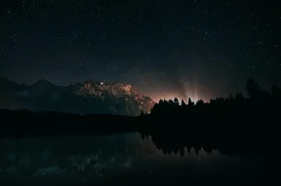 Stars fill the sky above a dark ridge, a faint glow behind the peaks and the whole scene mirrored in the water.