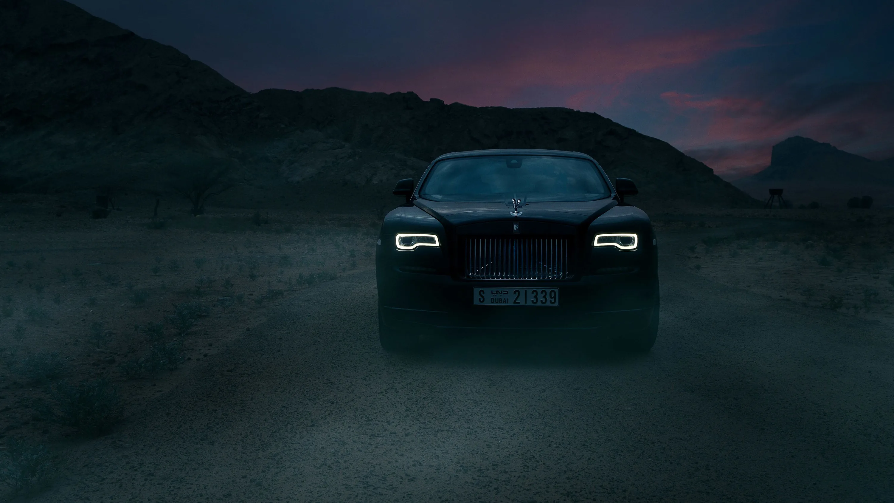 The coupe faces the camera on a dirt track, grille and daytime running lamps glowing faintly against a purple and pink dusk over dry hills.