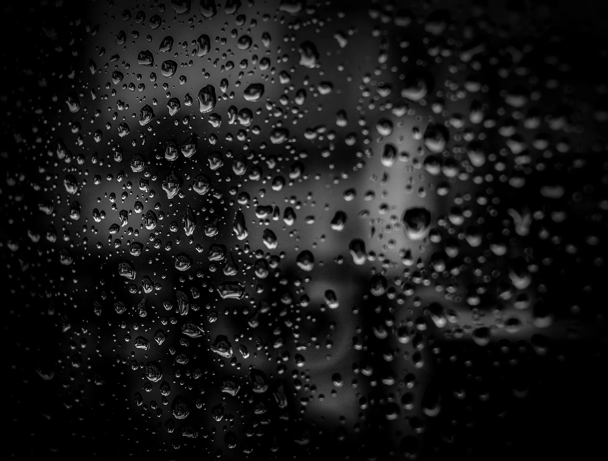Water beads cling to a pane of glass, city light behind them dissolved into soft grey blur and deep black.