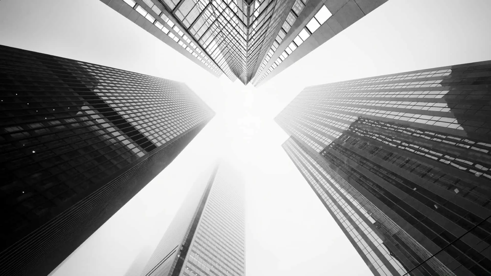 Four glass towers lean inward from the corners toward a blown-out white sky, shot straight up so the lines meet at the centre.