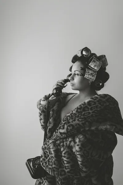 A studio portrait in monochrome — hair in metal rollers, a leopard print fur coat, one hand raised to the face against a plain pale wall.