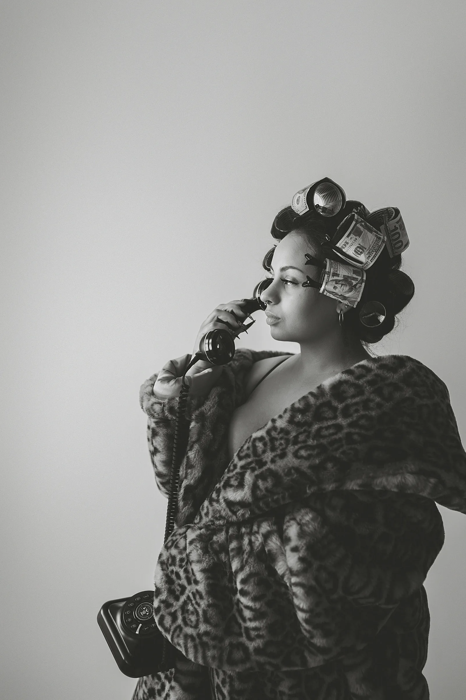 A studio portrait in monochrome — hair in metal rollers, a leopard print fur coat, one hand raised to the face against a plain pale wall.