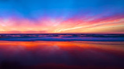 Motion-blurred clouds sweep in pink and white across a saturated blue sky, their colours smearing again through the surf and wet sand below.