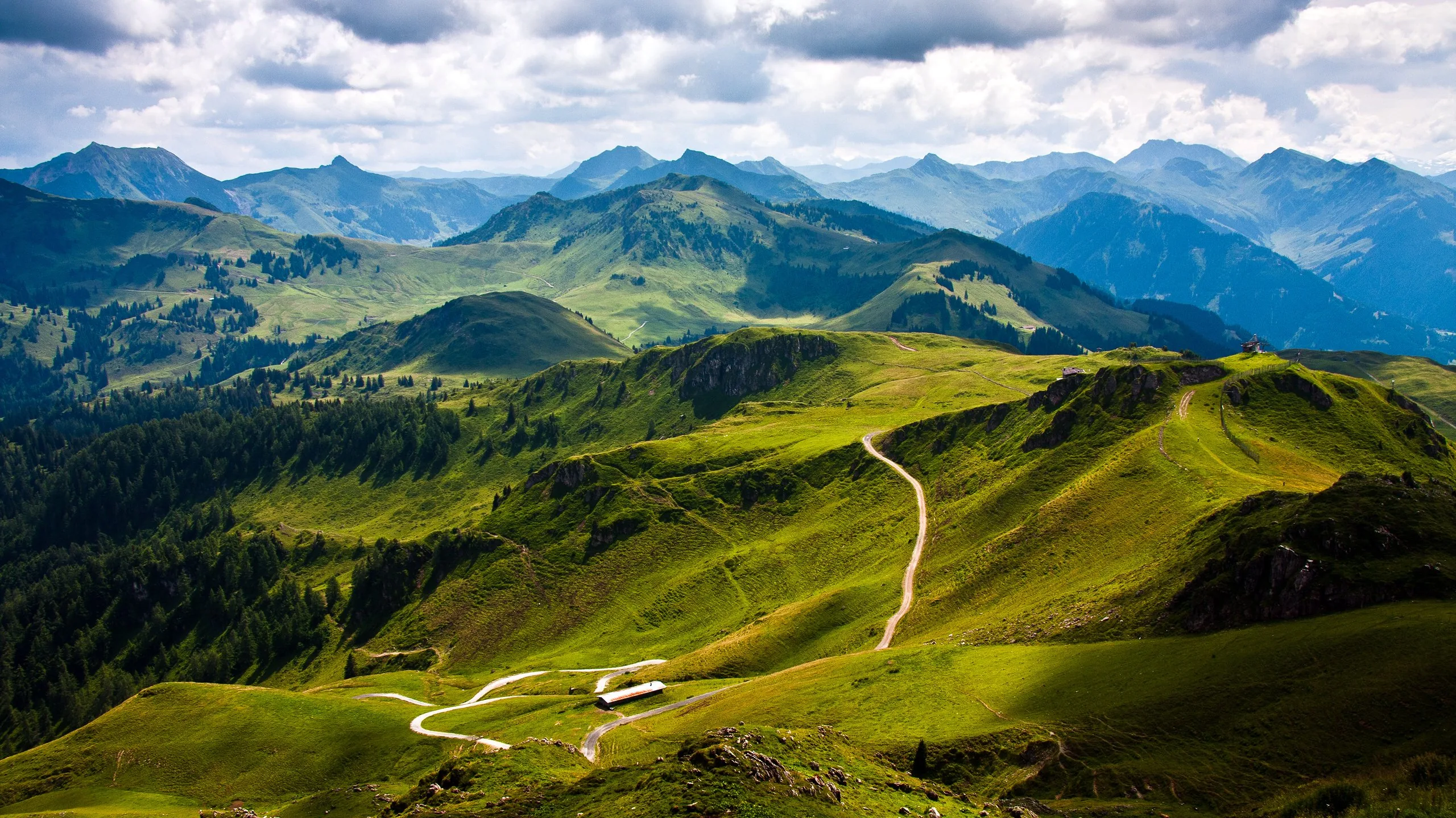 A dirt trail snakes along a grassy ridge past two small huts, layered green summits fading toward a cloud-scattered horizon.