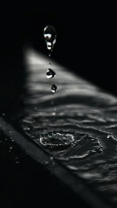A drop caught mid-fall above the ripple its predecessor left, shot in close monochrome with light raking across from the right.