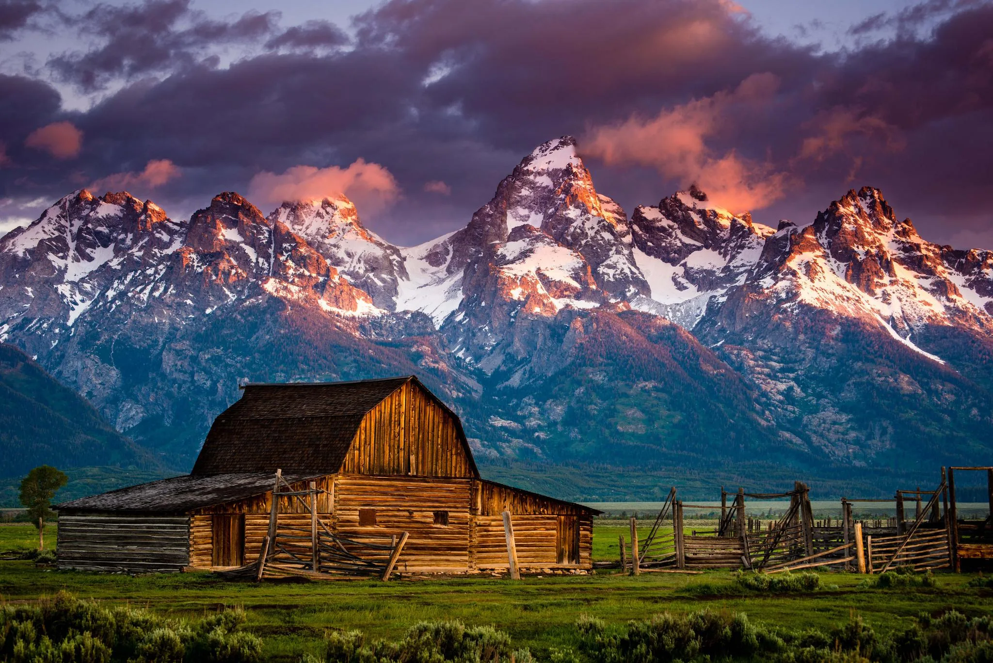 A weathered wooden barn sits in green pasture as pink sunrise clouds sweep over the snow-lined Teton range, the barn anchoring the lower centre.