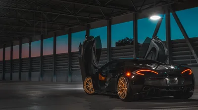Both doors swing skyward inside an empty warehouse, gold wheels and orange brake calipers glowing while teal and pink dusk shows through the window band.