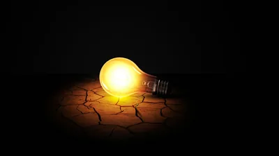 A lit bulb lies on parched, cracked ground, its yellow glow pooling along the bottom edge while the upper two-thirds stays solid black for a lock-screen clock.