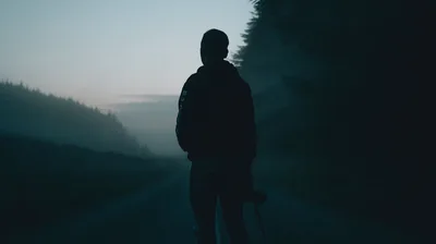 The road ahead dissolves into blue-grey fog past a man carrying something at his side, with the darkest pines stacked along the right edge.