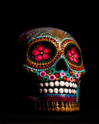 A Day of the Dead skull covered in coloured beadwork, flowers set into both eye sockets, lit from the left against black.