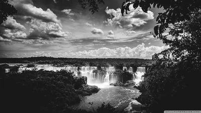 A long curtain of waterfalls drops into a river gorge, spray rising white against dark jungle and piled cumulus.