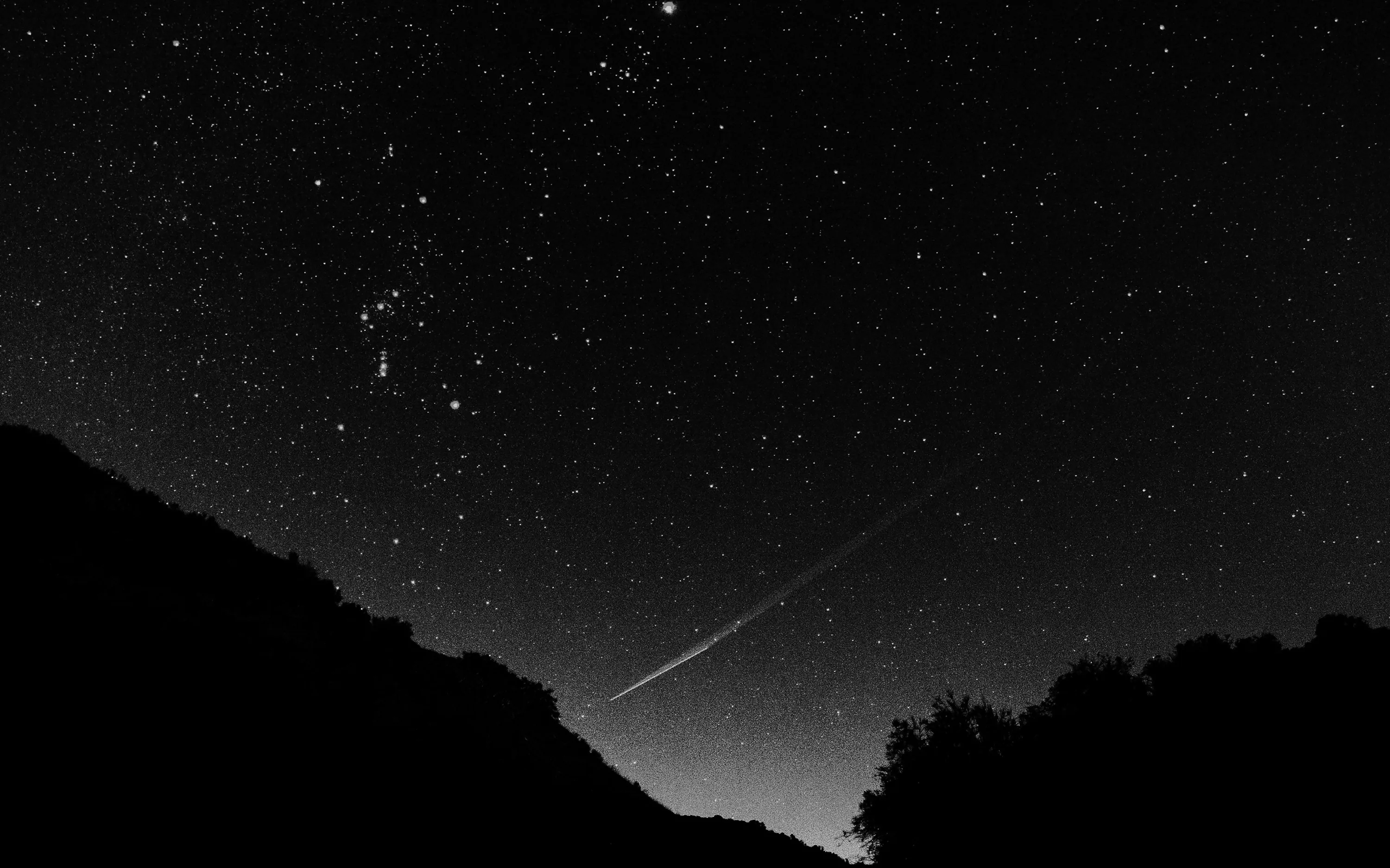 A meteor streaks down toward a black hilltop silhouette while a bright constellation sits in the upper left of the monochrome sky.