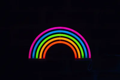 A rainbow bent from coloured neon tubes glows against a black brick wall, its light reaching only the bricks closest to the arc.
