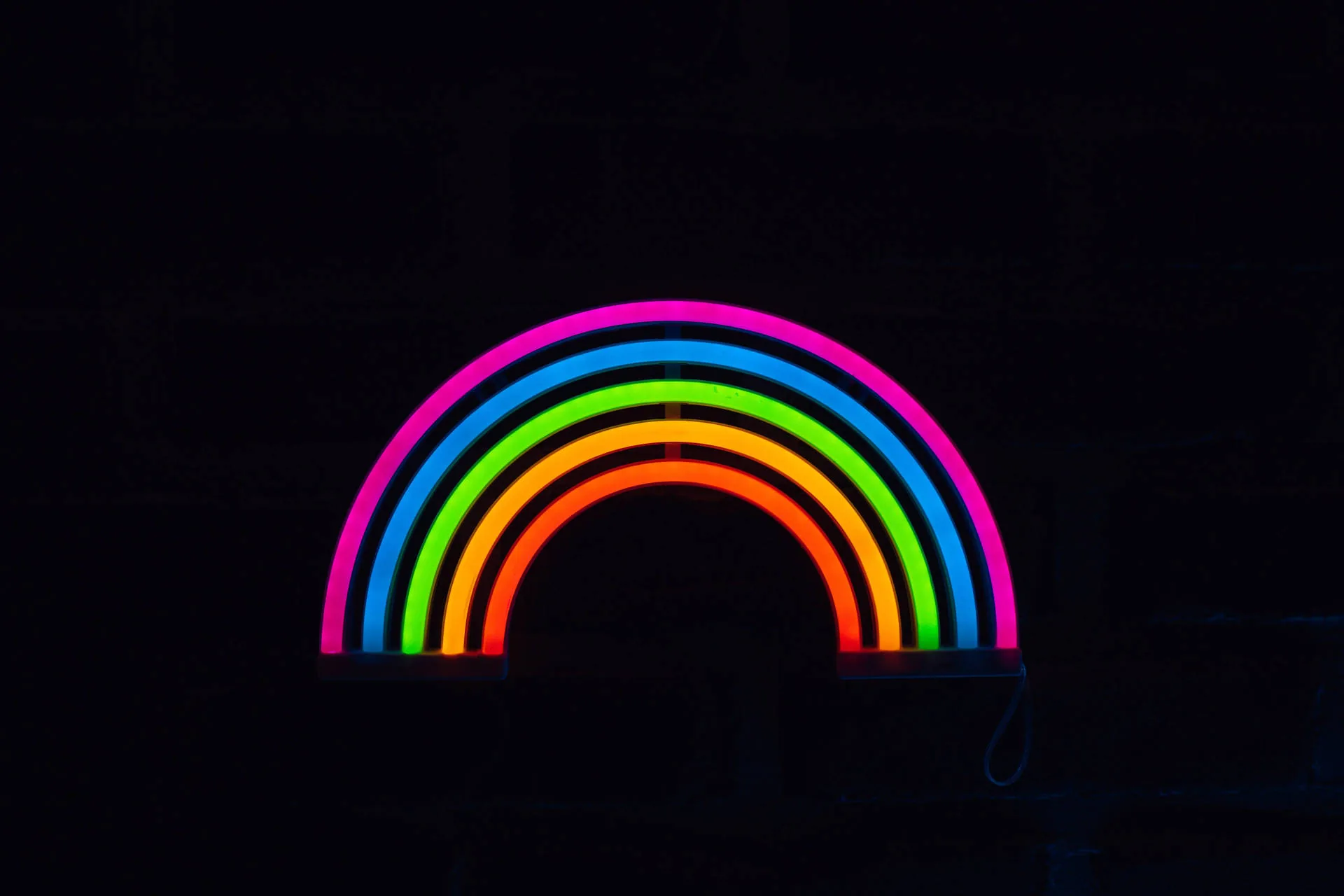 A rainbow bent from coloured neon tubes glows against a black brick wall, its light reaching only the bricks closest to the arc.