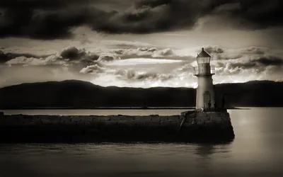A small lighthouse stands at the end of a stone breakwater in sepia monochrome, storm clouds gathering top-left while the water on the right stays calm and dark.