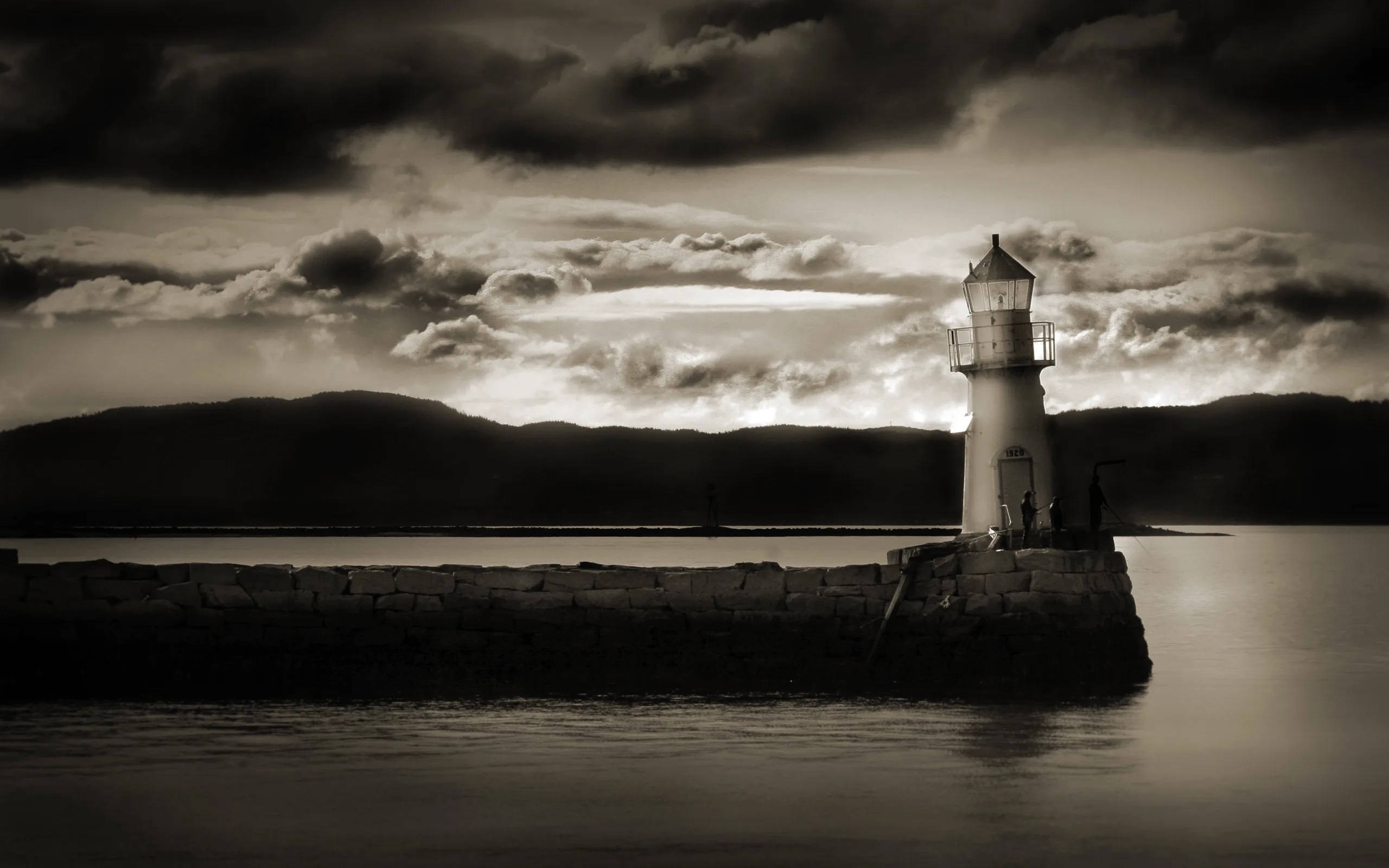 A small lighthouse stands at the end of a stone breakwater in sepia monochrome, storm clouds gathering top-left while the water on the right stays calm and dark.