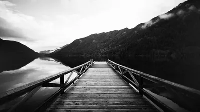 A weathered pier runs straight out over glassy water toward a forested ridge, mist hanging along the slope on the right.