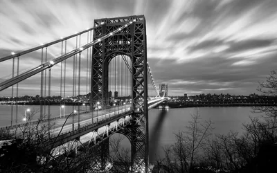 A steel suspension bridge crosses the river under streaked long-exposure cloud, its cables and deck lights picked out against a pale sky.