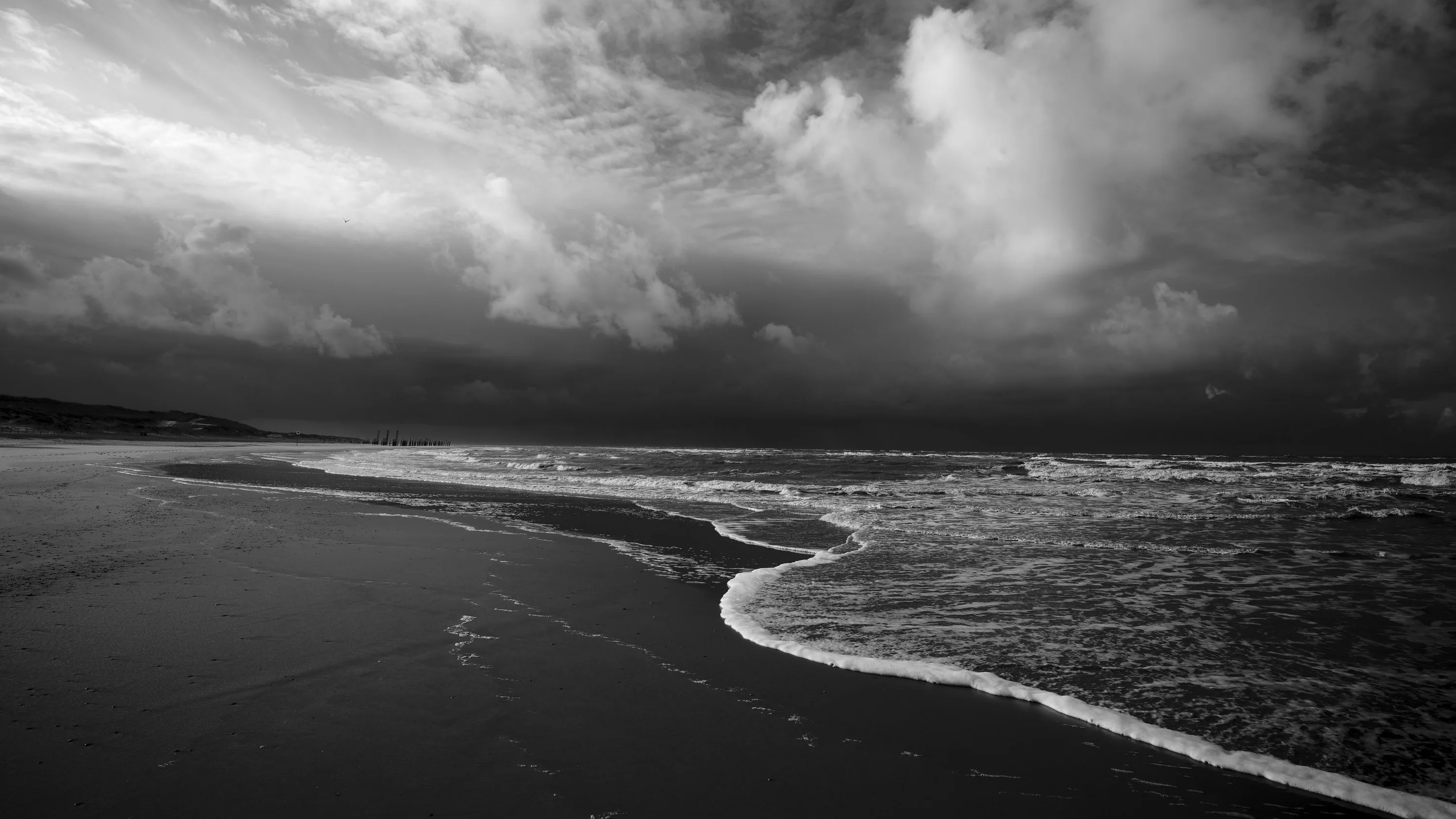 A curling wave runs up wet sand under a bank of storm cloud, the monochrome contrast strongest where the surf turns white.