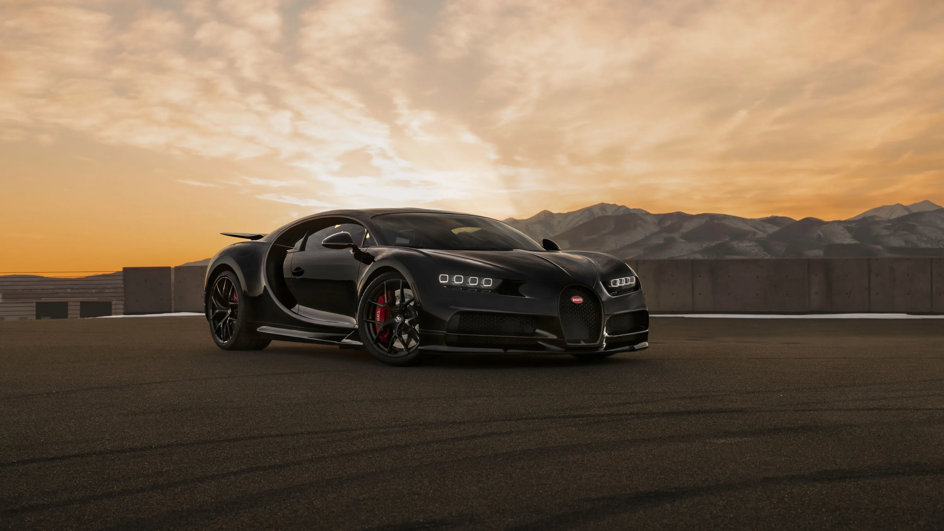 The Chiron rolls across bare tarmac as the sun drops behind a mountain ridge, orange sky filling the top half and red calipers showing through gold wheels.