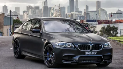 A satin black BMW sedan parks nose to the right on a city street, glass towers rising behind its roofline while soft daylight leaves the paint almost matte.