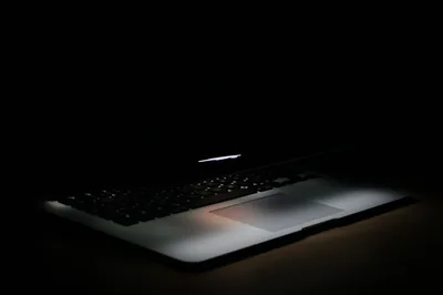 A laptop sits half in shadow with a thin line of light along the screen edge and the trackpad catching a glow.