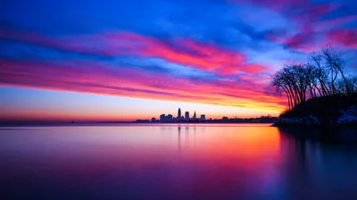 Pink cloud streaks cross a deep blue sky above a tiny distant skyline, bare trees on the right and glassy water keeping the rest of the frame quiet.