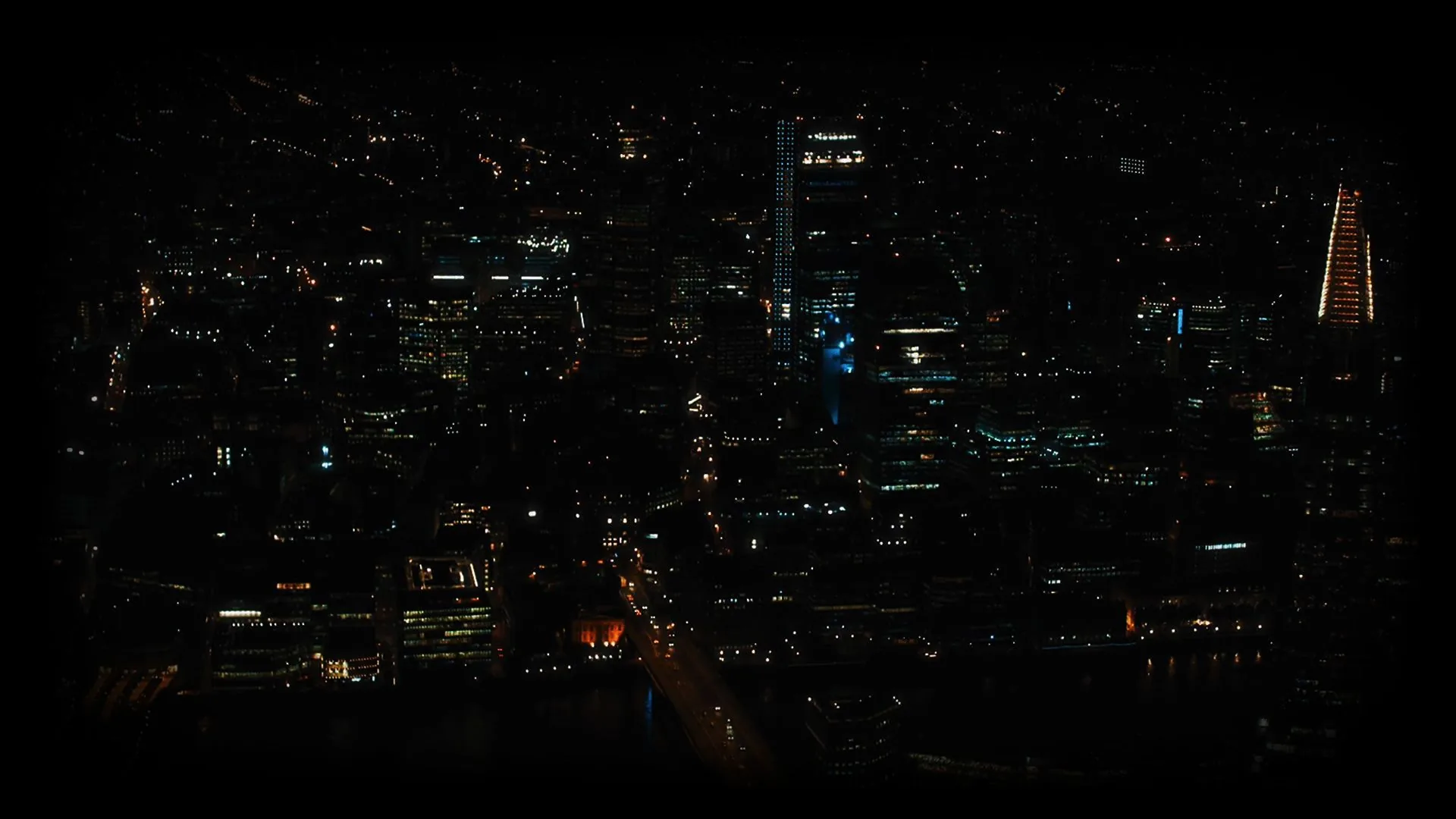Office towers and lit streets spread out below in a wide letterboxed frame, most of the grid dim with a few blue-lit facades standing out.
