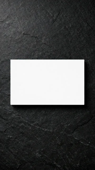 A blank white card lies flat and slightly above centre on rough black slate, its edge throwing a thin shadow down and to the right.