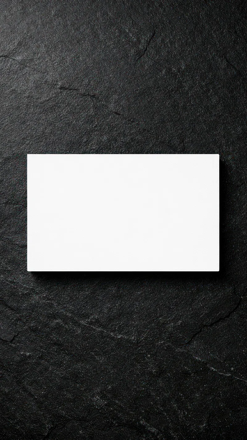 A blank white card lies flat and slightly above centre on rough black slate, its edge throwing a thin shadow down and to the right.