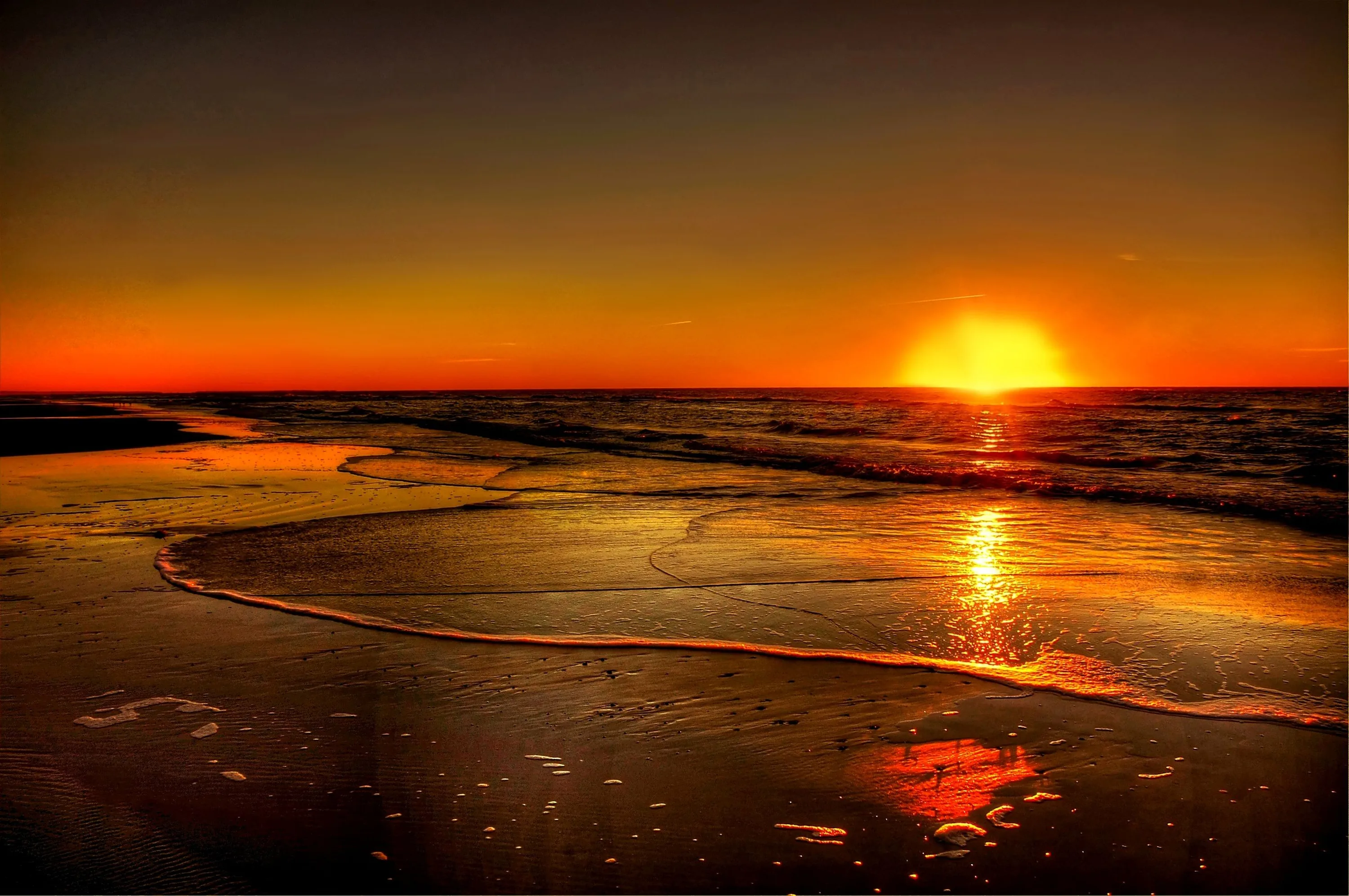 The last of the sun sits on the horizon at right, a curve of foam catching its glow while the rest of the beach falls to red-black — dark enough for OLED screens.
