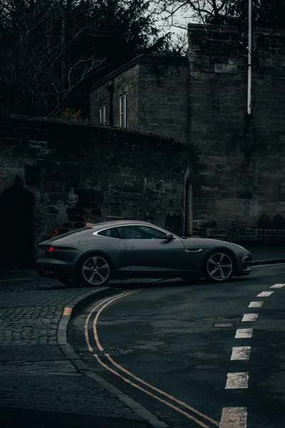 Dusk shot down a narrow lane: a dark coupe follows the double yellow line past an old stone wall, the cobbled kerb and bare trees kept in shadow.
