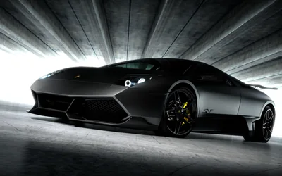 The car faces the camera head-on inside a ribbed concrete tunnel, daylight flaring in from both sides so the matte bodywork reads as one long shadow with yellow calipers behind the rims.