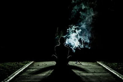 Flash-lit rails run out of the bottom corners toward a seated figure exhaling smoke to the right, the surrounding night pressing in as pure black.