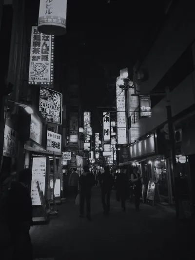 Stacked signboards glow down both sides of a narrow night alley as small figures walk the centre, the brightest lights clustered in the upper left.