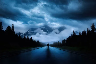 Rain-slicked and empty, the highway runs straight toward cloud-wrapped peaks with a walker at its centre line, blue storm light washing the whole scene.