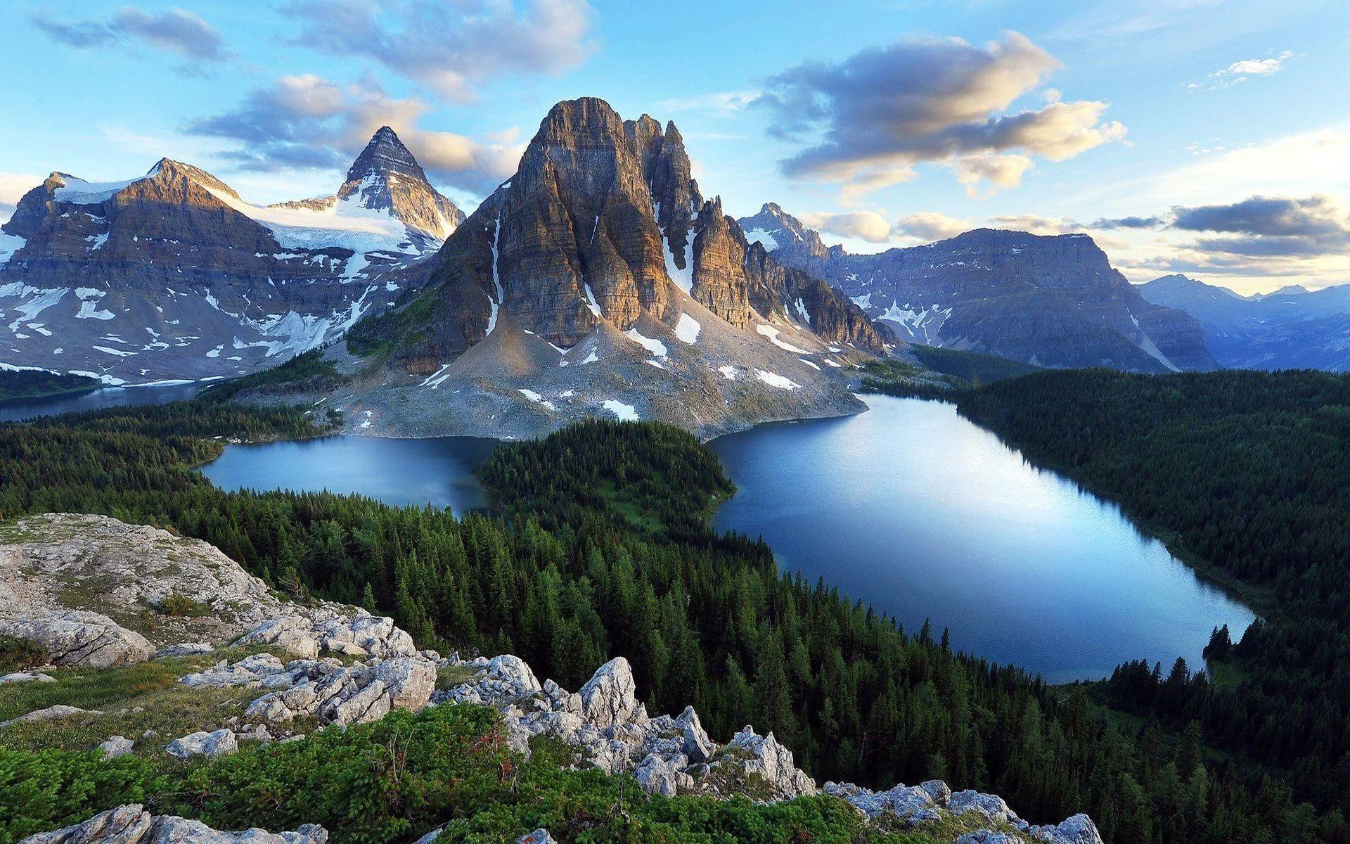 A craggy tower and its snowy neighbour stand over two deep blue lakes and unbroken spruce forest, seen from a boulder-strewn ridge at golden hour.