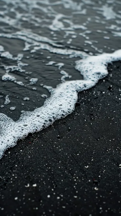 A lace of surf crosses wet volcanic sand from the upper left, droplets scattered over the dark grain that fills the lower half.