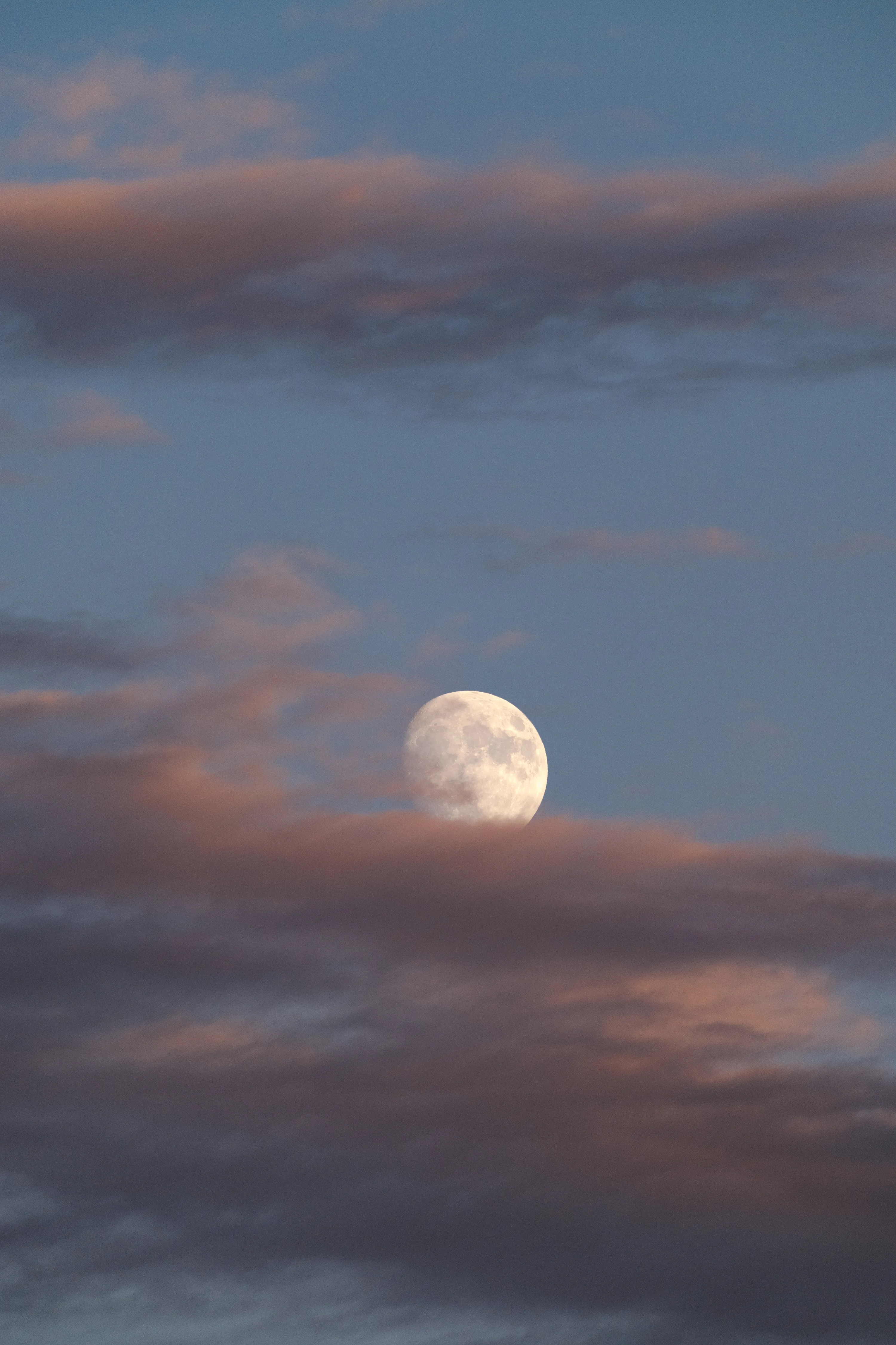 A waxing gibbous moon sits mid-frame between layers of pink-lit evening cloud, the surrounding blue sky staying plain enough for app icons.