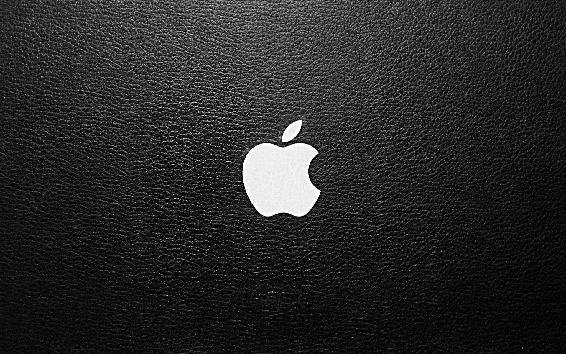 The Apple logo sits in white on grained black leather, slightly right of centre, lit softly from above so the pores stay visible.
