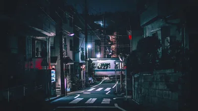 A quiet alley runs between low buildings after dark, a crosswalk in the foreground and one bright shopfront glowing at the far end.