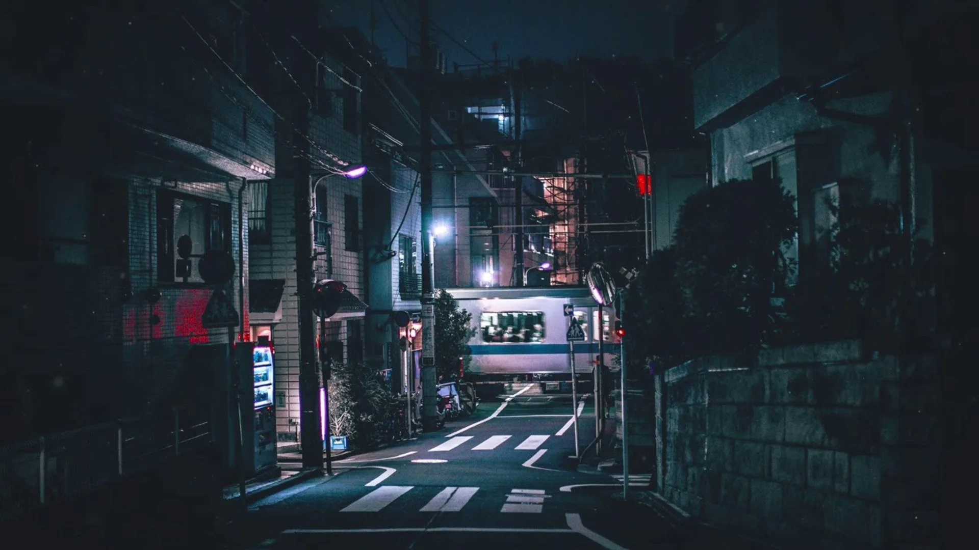A quiet alley runs between low buildings after dark, a crosswalk in the foreground and one bright shopfront glowing at the far end.