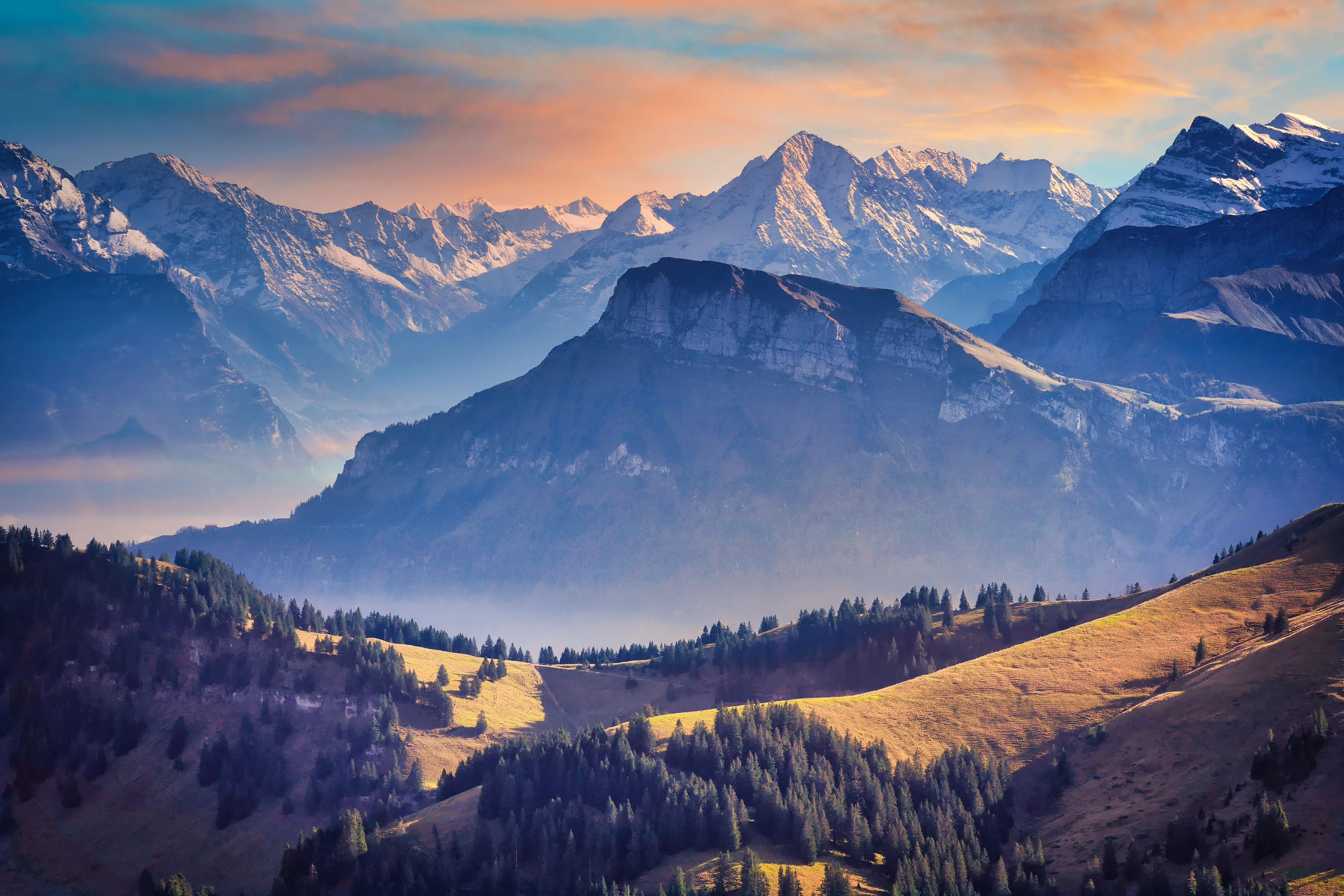 Ridge after ridge recedes into blue haze toward snow peaks under a peach sky, warm light grazing the pastured hills in the foreground.