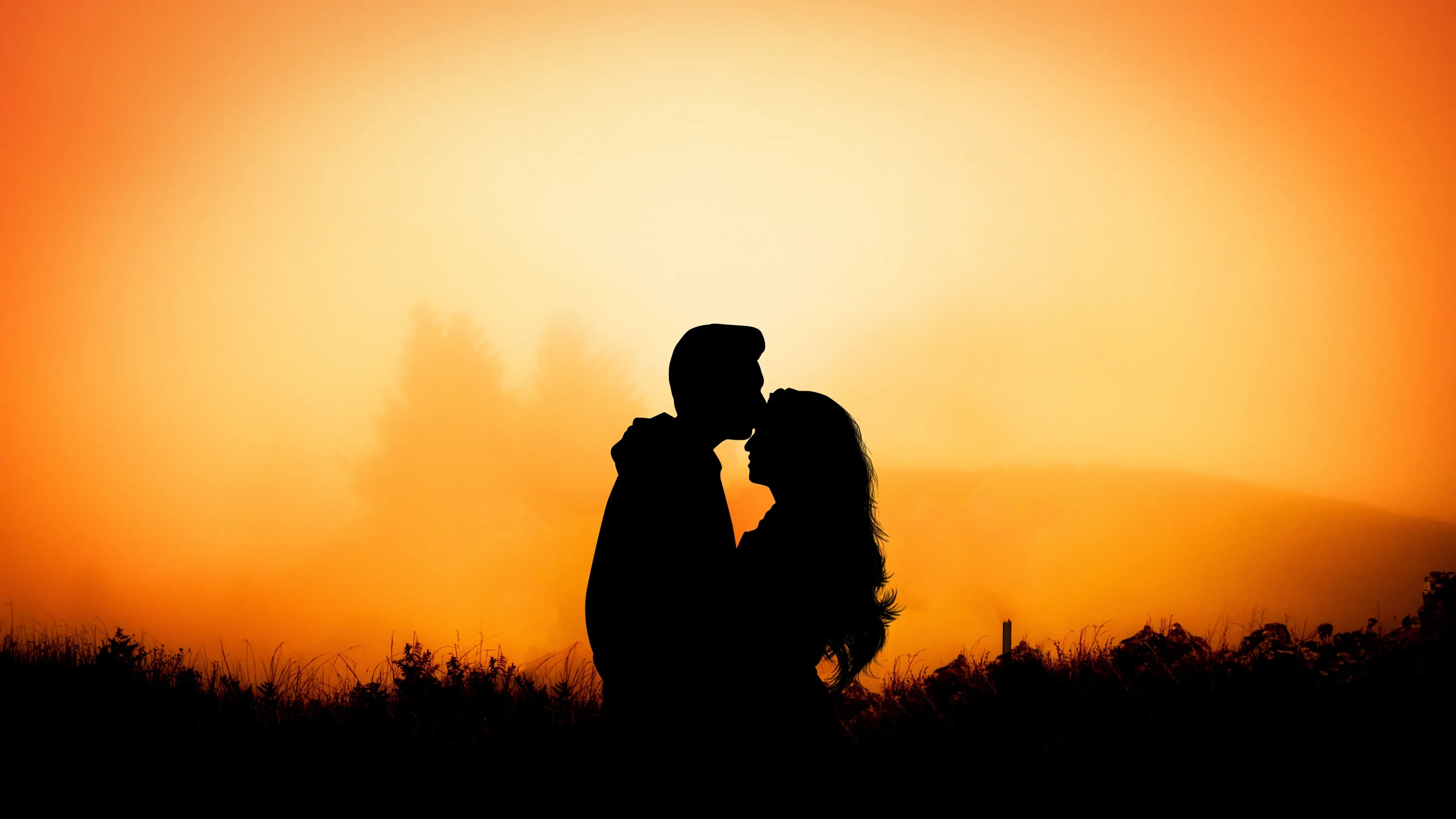 Two silhouettes meet for a kiss against a field of burnt orange haze, only the dark line of grass and low trees breaking the bottom of the frame.