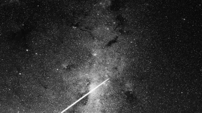 A bright streak cuts diagonally through the dusty band of the Milky Way, dark dust lanes breaking up the crowded stars behind it.