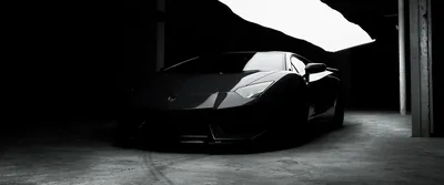 Ultrawide crop of a Lamborghini in an unlit parking structure, hard daylight from a doorway cutting one bright wedge across the roof and windscreen.